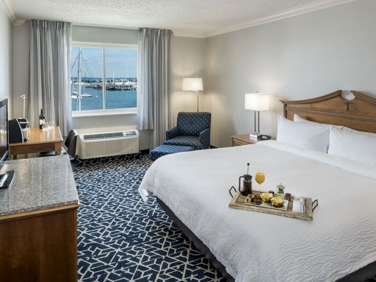 A hotel room with a large bed, tray with drinks, TV, armchair, and window with a harbor view under soft lighting completes the scene.