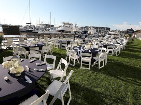 An outdoor event setup features round tables with white chairs, floral centerpieces, and a marina with boats in the background.