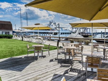 The image shows a sunny marina scene with wooden decking, tables and chairs, large yellow umbrellas, boats, and a grassy area.