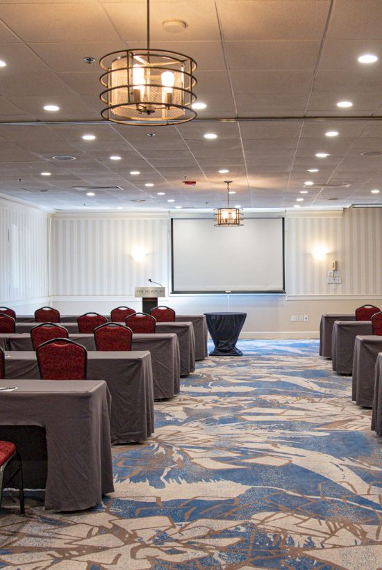 A conference room with rows of tables and chairs, projector screen, and patterned carpet, ready for a meeting or presentation.