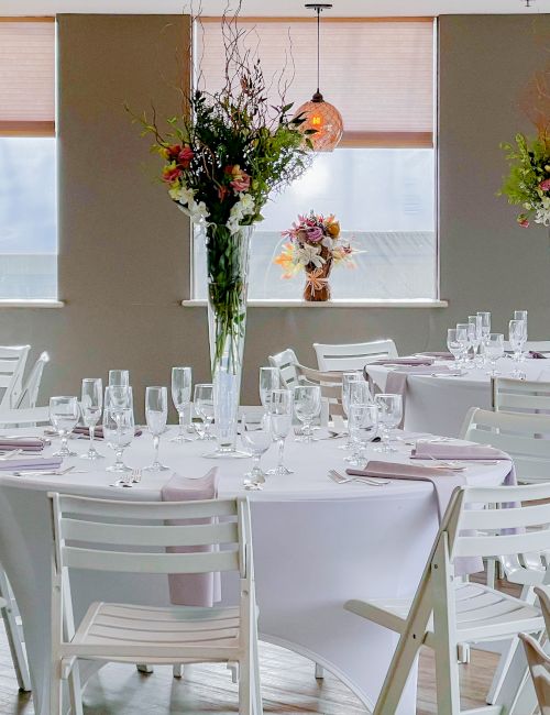 A decorated event space with round tables, white chairs, floral centerpieces, and soft lighting from hanging lamps by the windows.