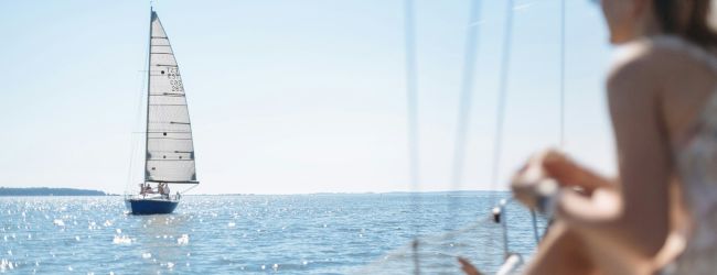 A person enjoys a sunny day on a boat, watching a sailboat on the calm water.