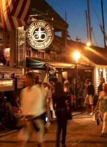 A lively evening street scene with people walking, illuminated shops, and an American flag prominently displayed.