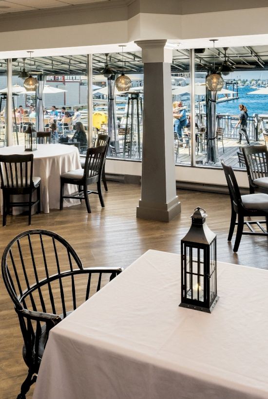 A restaurant interior with black chairs, white tablecloths, and lanterns, overlooking a waterfront patio through large windows, is shown.