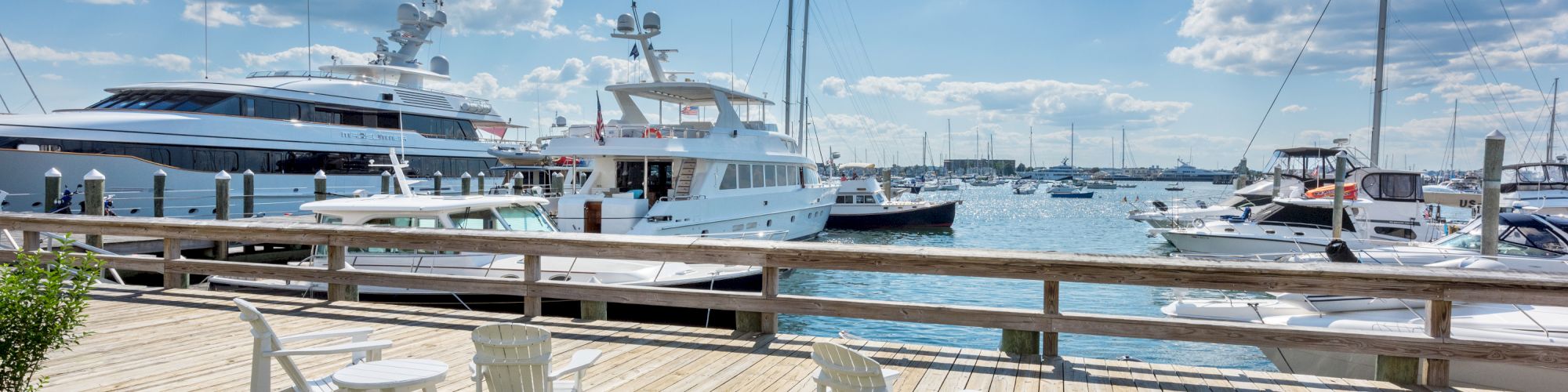 A scenic view of a marina with yachts docked, a wooden deck featuring Adirondack chairs and tables, under a cloudy blue sky.