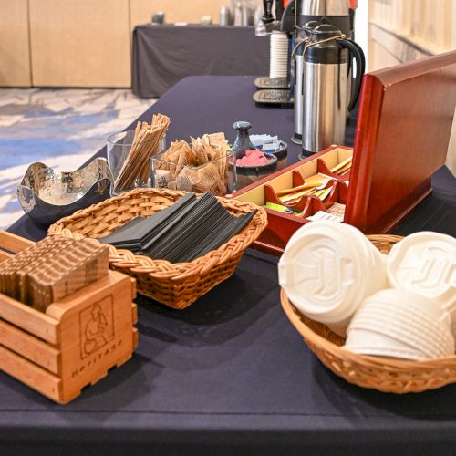 A coffee station with cups, stirrers, a coffee dispenser, and various condiments is set up on a table.