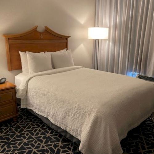 The image shows a tidy hotel room with a neatly made bed, two bedside lamps, a wooden headboard, a side table, and curtains.