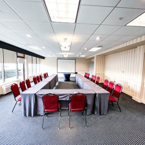 The image shows a conference room with U-shaped table setup, red chairs, projector screen, and large windows on one side of the room.