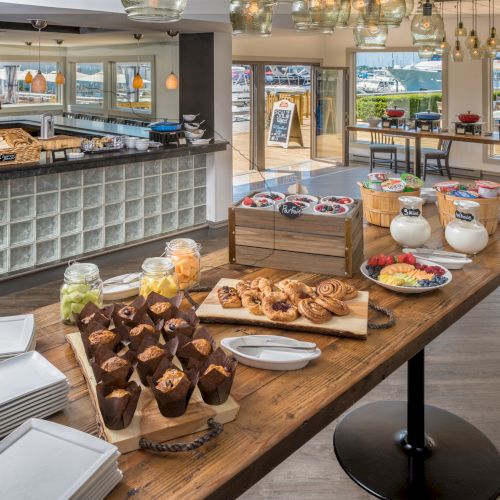 The image shows a buffet with muffins, pastries, yogurts, and fruits on a wooden table in a modern dining area with glass accents.