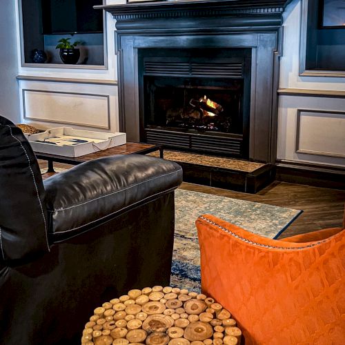 A cozy living room with a fireplace, black couch, orange chair, wooden coffee table, and framed pictures above the mantel.