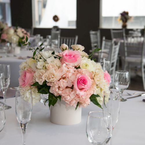 A decorated table with pink and white flower centerpiece, surrounded by empty wine glasses, in a formal setting with other similar tables.