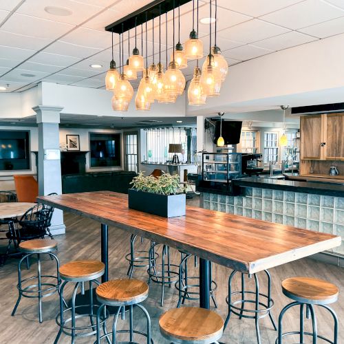 A modern cafe with wooden tables, hanging lights, and industrial-style stools; features a central high table with a small plant arrangement.