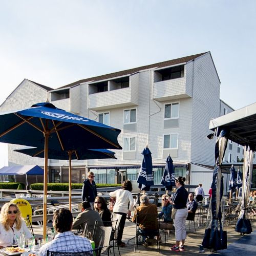 People dining outdoors by a waterfront, with a modern white building and boats in the background, under umbrellas.