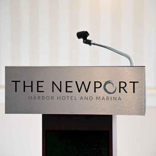 A podium with a microphone is shown, labeled "THE NEWPORT Harbor Hotel and Marina," set against a striped background.