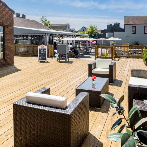 A wooden deck with wicker furniture, glass-topped tables, potted plants, and a view of a patio area with more seating and umbrellas.