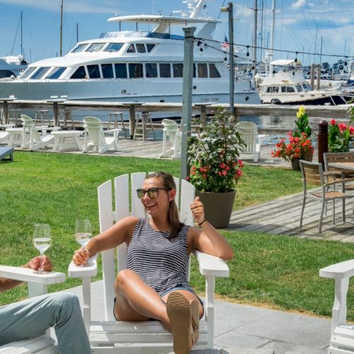 Two people relax on Adirondack chairs with drinks, overlooking a marina with yachts and sailboats. Bright, sunny, and serene atmosphere.