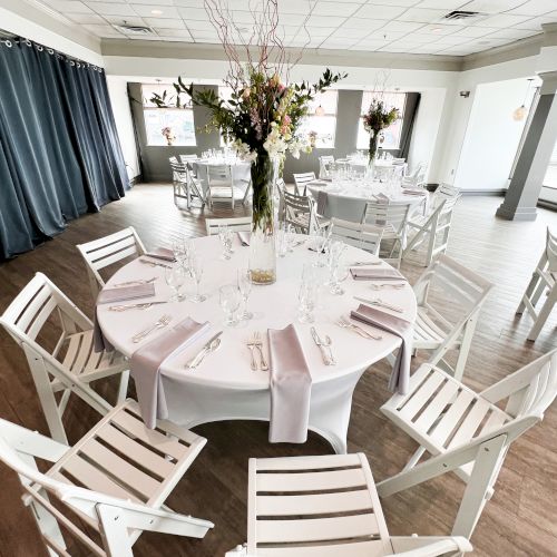 An elegantly set round table with white chairs, adorned with a tall floral centerpiece, is in a bright room with large windows and curtains.