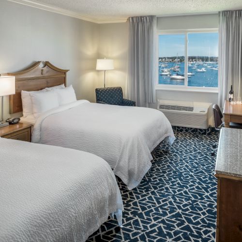 The image shows a hotel room with two beds, a desk, a TV, and a window overlooking a marina with boats.