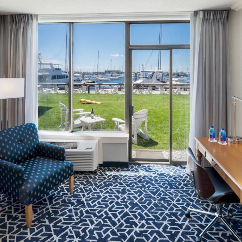 A hotel room with a chair, desk, and harbor view through large windows. There are boats, a lawn, and a couple of chairs outside.