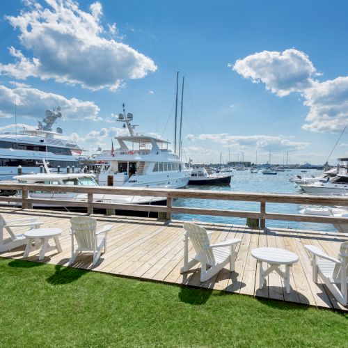 The image shows a marina with yachts, a wooden deck with chairs and tables, and a grass area under a partly cloudy blue sky.