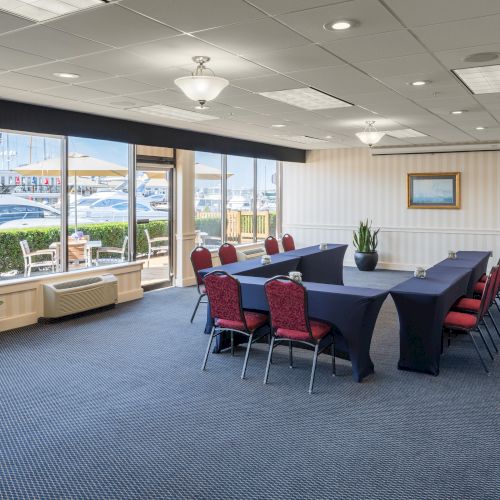 A conference room with a U-shaped table setup and red chairs overlooks a marina through large windows. The room is bright and spacious.