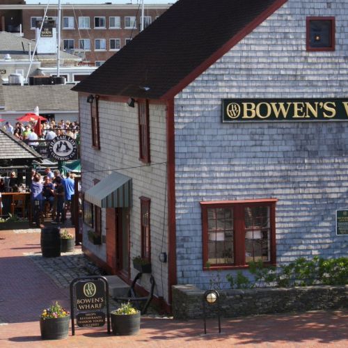 The image shows Bowen's Wharf with a bustling group of people gathered near a building, set against a backdrop of marina and boats.