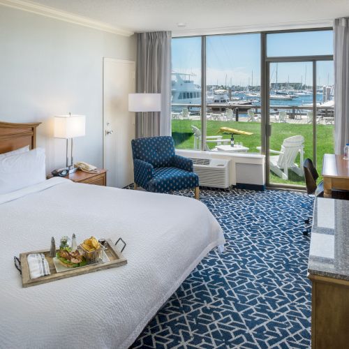 A hotel room with a king-sized bed, tray with food, TV, patterned carpet, blue chair, and a view of a marina outside the window.