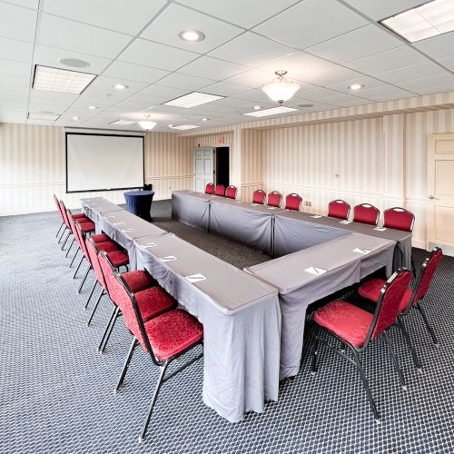 A conference room with a U-shaped table setup, red chairs, a projector screen, and striped wallpaper. It's well-lit with ceiling lights.