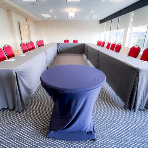 A conference room setup with U-shaped tables and red chairs, featuring a covered round table at the center, on a carpeted floor.