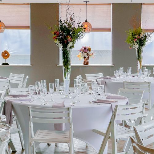 The image shows a decorated dining area with round tables, white chairs, floral centerpieces, and hanging lights in a bright room.