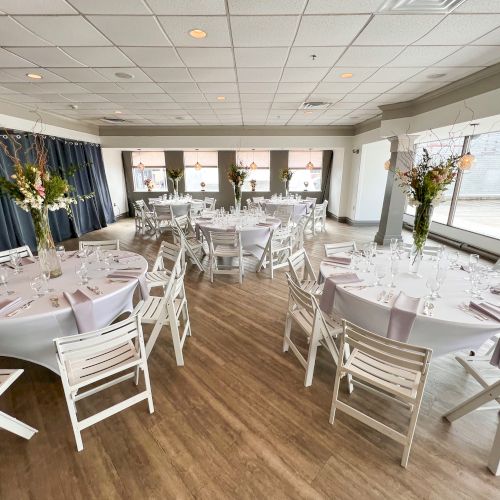 The image shows a decorated event space with round tables, white chairs, and floral centerpieces in a bright room.