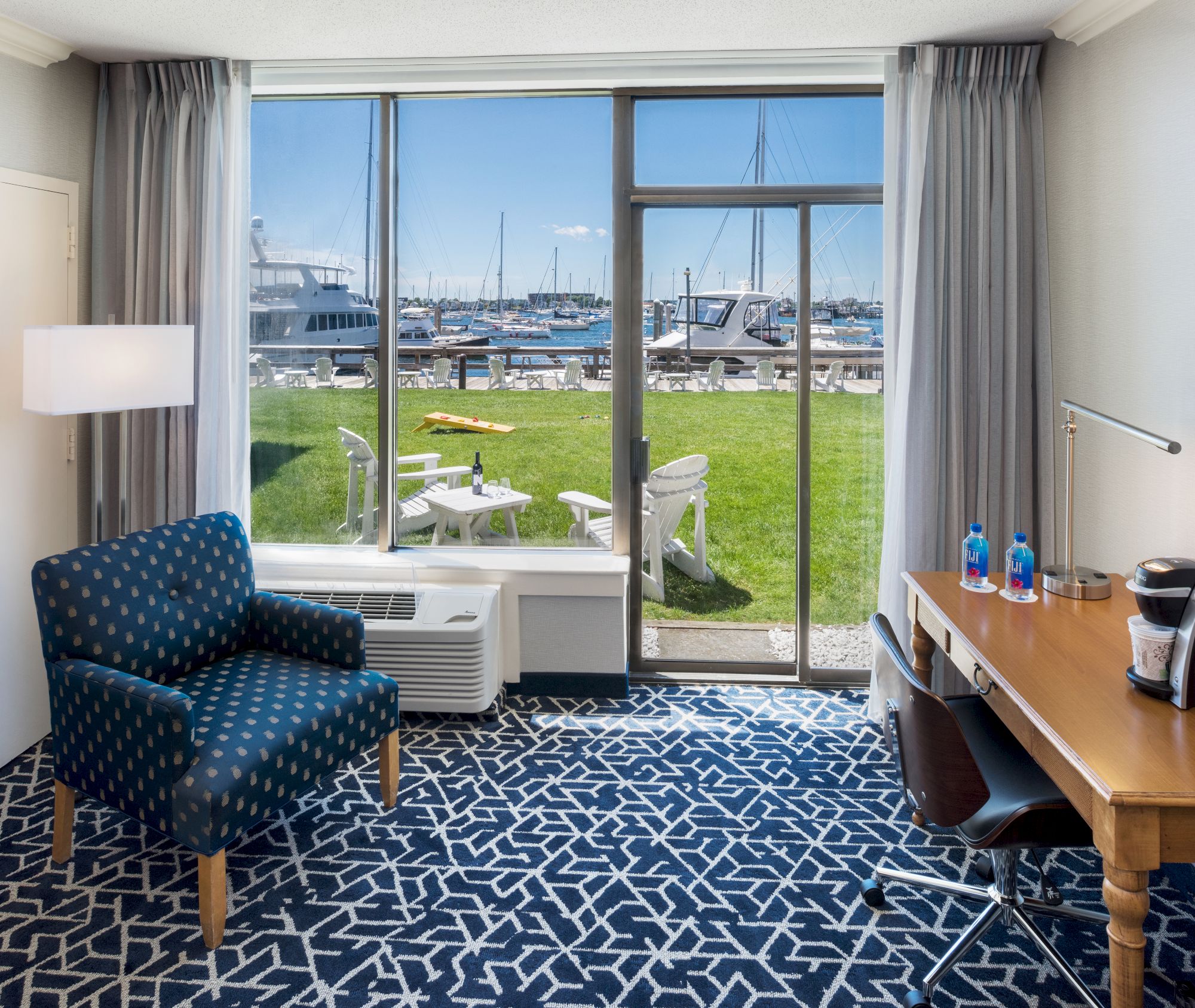 A hotel room with a patterned carpet, chair, desk, and a view of boats and water through large windows, leads to a grassy area with chairs.