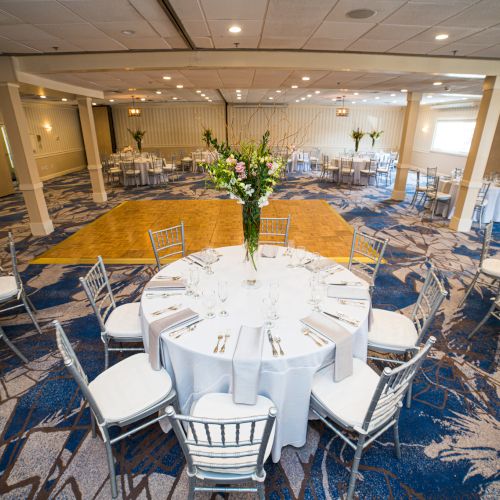 A decorated banquet hall with round tables, white tablecloths, silver chairs, and floral centerpieces on a patterned carpet floor.