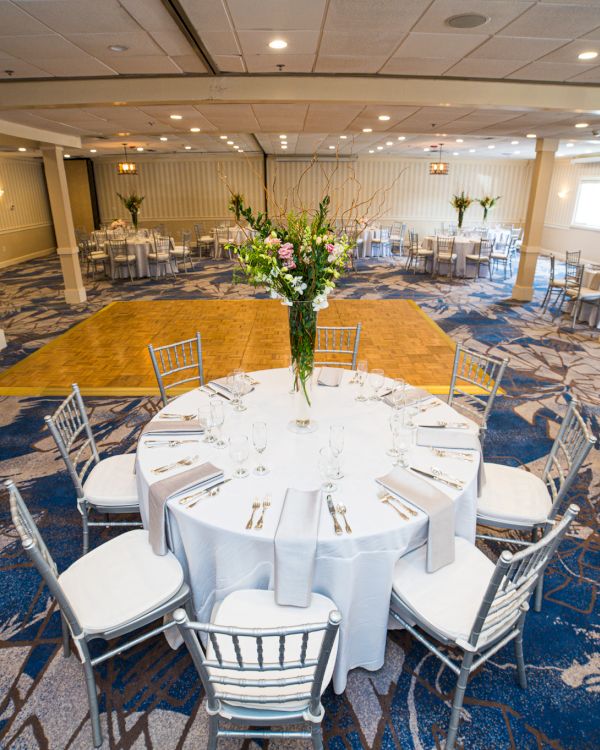 A decorated banquet hall with round tables, white tablecloths, silver chairs, and floral centerpieces on a patterned carpet floor.