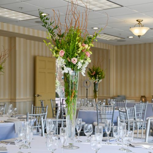 An elegant banquet room with round tables, silver chairs, and tall floral centerpieces set for a formal event or wedding reception.