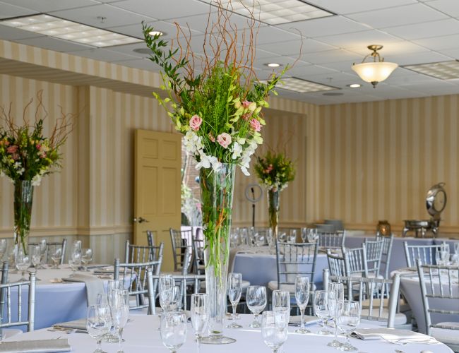 An elegant banquet room with round tables, silver chairs, and tall floral centerpieces set for a formal event or wedding reception.