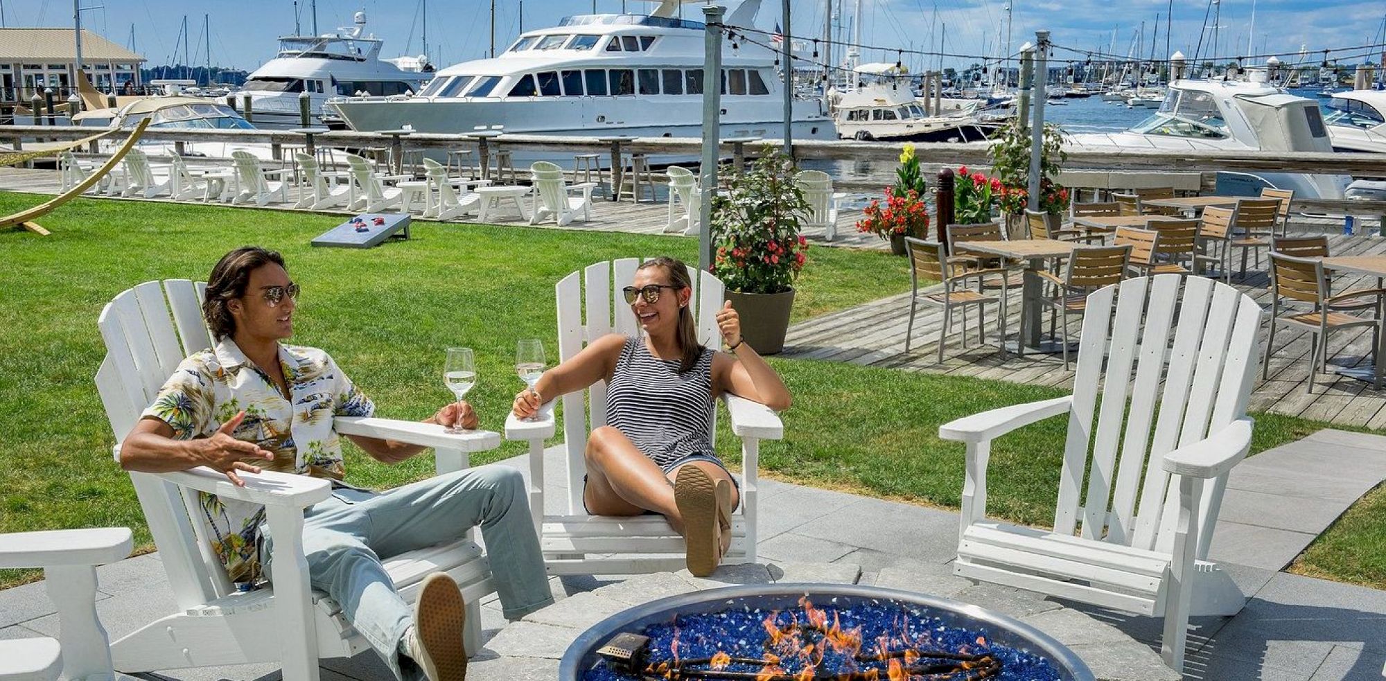 Two people relaxing in Adirondack chairs by a waterfront with yachts, enjoying drinks by a fire pit on a sunny day.