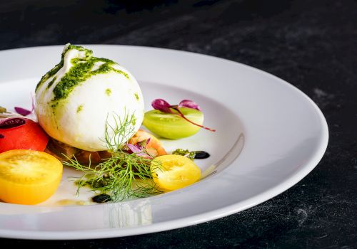 The image shows a plate of burrata cheese with colorful cherry tomatoes, herbs, and a drizzle of green sauce on a white dish.