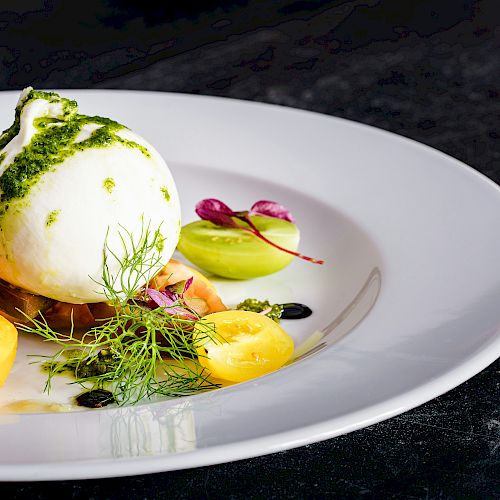 The image shows a plate of burrata cheese with colorful cherry tomatoes, herbs, and a drizzle of green sauce on a white dish.