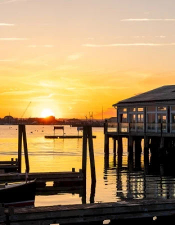 A serene sunset over a waterfront with two dockside buildings and calm waters, casting beautiful reflections in the scene.