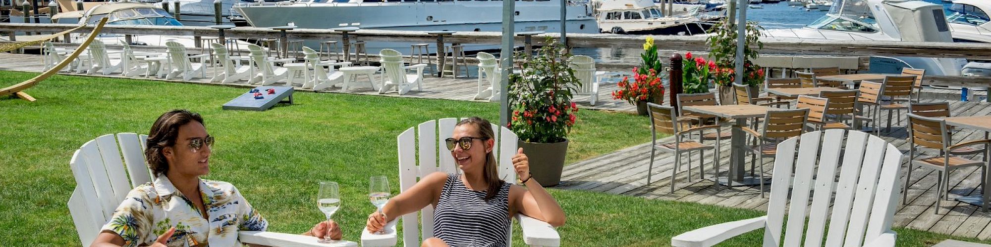 Two people relax in Adirondack chairs on a lawn by a marina, enjoying drinks under a sunny sky, with boats docked in the background.